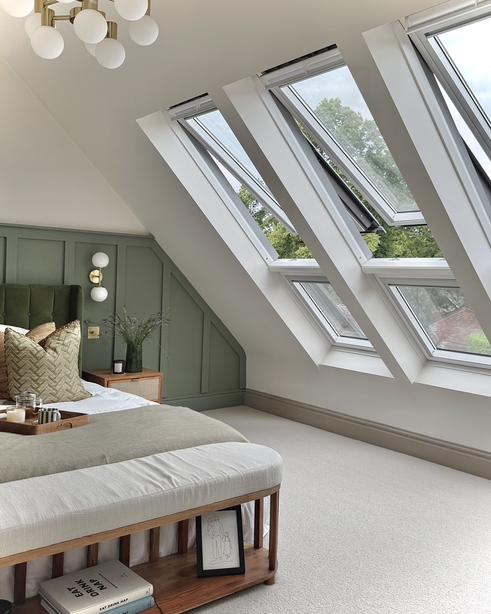 Modern bedroom with sage green wall panelling covering half of the wall from the bottom, with a large bed and three open, large pitched roof windows to the right
